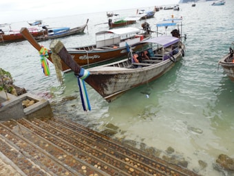 Wooden boats are docked along the shoreline in a serene body of water. Some boats are adorned with colorful ribbons tied around the bows. Steps lead down to the water, indicating easy access to the boats. The background shows more boats floating calmly on the water.