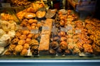 An assortment of pastries and baked goods displayed in a bakery window, including cookies, croissants, and cakes. Each item is labeled with a price tag. The pastries vary in shapes and sizes, with some topped with almonds and others drizzled with chocolate.