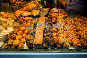 An assortment of pastries and baked goods displayed in a bakery window, including cookies, croissants, and cakes. Each item is labeled with a price tag. The pastries vary in shapes and sizes, with some topped with almonds and others drizzled with chocolate.