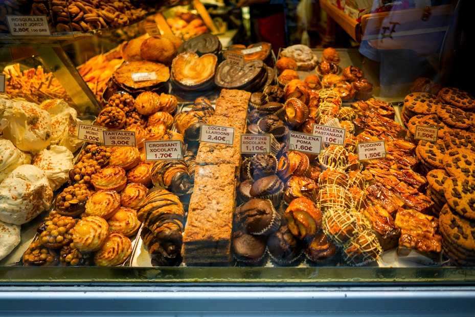 An assortment of pastries and baked goods displayed in a bakery window, including cookies, croissants, and cakes. Each item is labeled with a price tag. The pastries vary in shapes and sizes, with some topped with almonds and others drizzled with chocolate.