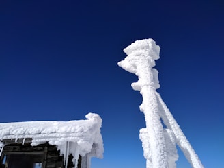 A structure is heavily covered in snow or ice against a clear blue sky. The buildup of ice creates thick, frosty layers on the wooden surface and a tall, narrow installation.