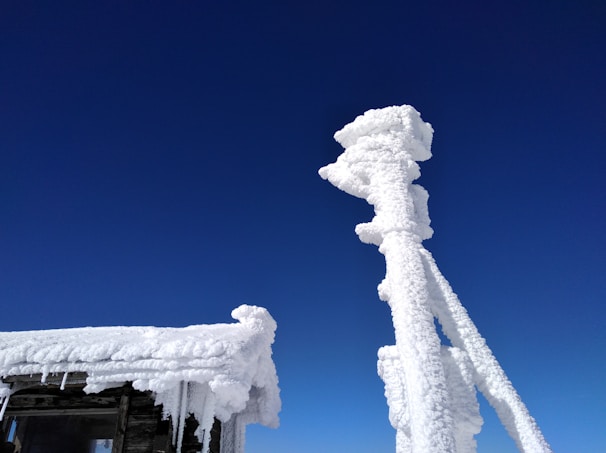 A structure is heavily covered in snow or ice against a clear blue sky. The buildup of ice creates thick, frosty layers on the wooden surface and a tall, narrow installation.