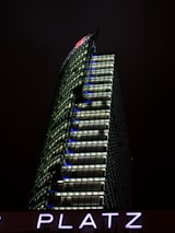 A modern skyscraper with a distinctive angled design is illuminated against a dark night sky. The building features numerous windows with bright lights, and the letters 'DB' are prominently displayed at the top. Below, the letters 'PLATZ' are visible, lit in a glowing purple hue.