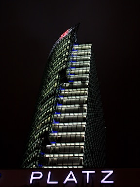 A modern skyscraper with a distinctive angled design is illuminated against a dark night sky. The building features numerous windows with bright lights, and the letters 'DB' are prominently displayed at the top. Below, the letters 'PLATZ' are visible, lit in a glowing purple hue.