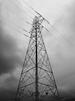 A tall metal electrical tower stands against a backdrop of overcast skies, with wires extending outwards. The structure's intricate lattice pattern is highlighted by the contrast with the cloudy sky.