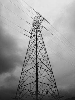 A tall metal electrical tower stands against a backdrop of overcast skies, with wires extending outwards. The structure's intricate lattice pattern is highlighted by the contrast with the cloudy sky.