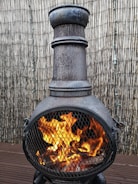 A metallic outdoor chiminea with a tall chimney and a spherical body, placed on a wooden deck. Bright orange flames are visible through the mesh covering, surrounded by a vertical bamboo fence in the background.