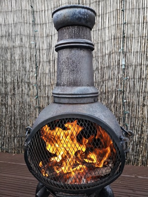 A metallic outdoor chiminea with a tall chimney and a spherical body, placed on a wooden deck. Bright orange flames are visible through the mesh covering, surrounded by a vertical bamboo fence in the background.