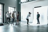 A photography studio scene featuring a photographer capturing a male subject who is posing on a white backdrop. Several people are observed behind the photographer, including a woman adjusting equipment and others casually standing around. The room is industrial-style with large windows and visible vents on the ceiling.