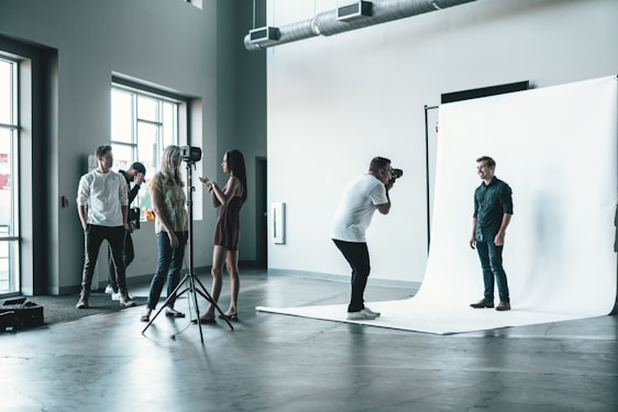 A photography studio scene featuring a photographer capturing a male subject who is posing on a white backdrop. Several people are observed behind the photographer, including a woman adjusting equipment and others casually standing around. The room is industrial-style with large windows and visible vents on the ceiling.