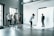 A photography studio scene featuring a photographer capturing a male subject who is posing on a white backdrop. Several people are observed behind the photographer, including a woman adjusting equipment and others casually standing around. The room is industrial-style with large windows and visible vents on the ceiling.