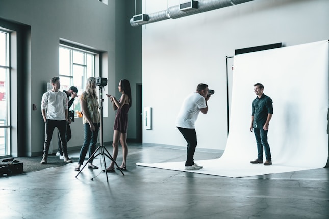 A photography studio scene featuring a photographer capturing a male subject who is posing on a white backdrop. Several people are observed behind the photographer, including a woman adjusting equipment and others casually standing around. The room is industrial-style with large windows and visible vents on the ceiling.