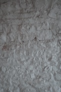 A textured surface of a rough, uneven gray wall. The wall's surface is characterized by a stucco finish with visible bumps and ridges, creating a rustic and tactile appearance.