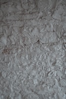 A textured surface of a rough, uneven gray wall. The wall's surface is characterized by a stucco finish with visible bumps and ridges, creating a rustic and tactile appearance.