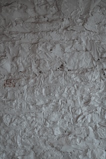 A textured surface of a rough, uneven gray wall. The wall's surface is characterized by a stucco finish with visible bumps and ridges, creating a rustic and tactile appearance.