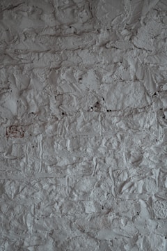 A textured surface of a rough, uneven gray wall. The wall's surface is characterized by a stucco finish with visible bumps and ridges, creating a rustic and tactile appearance.