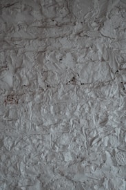 A textured surface of a rough, uneven gray wall. The wall