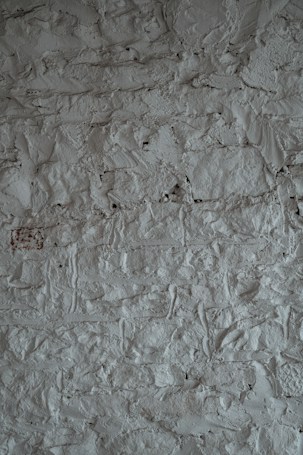A textured surface of a rough, uneven gray wall. The wall's surface is characterized by a stucco finish with visible bumps and ridges, creating a rustic and tactile appearance.