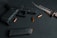 A black handgun is laid on a dark, textured surface alongside a serrated knife and several brass-colored bullets. The arrangement of these items suggests a focus on weaponry and ammunition.