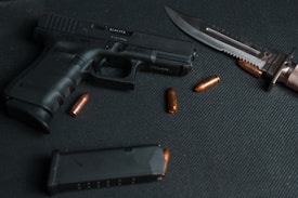 A black handgun is laid on a dark, textured surface alongside a serrated knife and several brass-colored bullets. The arrangement of these items suggests a focus on weaponry and ammunition.