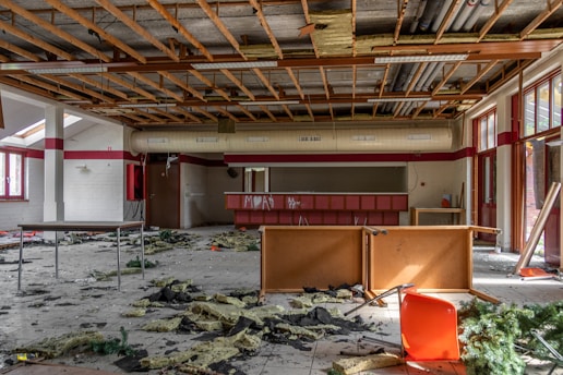 A room with a partially destroyed ceiling, exposing wooden beams and insulation. Debris is scattered across the tiled floor, and there are remnants of furniture including tables and a red chair. Windows are present, allowing natural light to enter, and graffiti is visible on a counter.