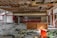 A room with a partially destroyed ceiling, exposing wooden beams and insulation. Debris is scattered across the tiled floor, and there are remnants of furniture including tables and a red chair. Windows are present, allowing natural light to enter, and graffiti is visible on a counter.