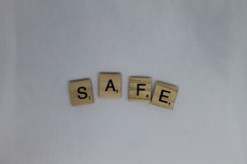 Four Scrabble tiles spelling the word SAFE are arranged horizontally on a textured white fabric background. Each tile has a letter with a corresponding point value in the lower right corner.