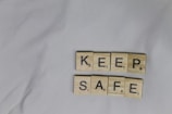 Wooden tiles with letters spelling out 'KEEP SAFE' are arranged in two rows on a light-colored, slightly wrinkled fabric background.