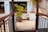 An outdoor area featuring a pathway lined with dark wooden railings leading to a patio with potted plants. The patio shows tiled flooring and steps that ascend on both sides. There is a large potted palm and smaller plants placed around, giving the space a tropical feel.