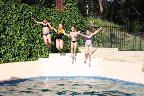 Four children in swimsuits are joyfully jumping into a swimming pool from the ledge. The background features lush green foliage and a fence, with sunlight casting clear shadows, creating a lively and vibrant scene.