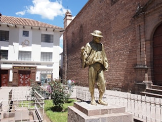 A bronze statue of a person wearing a traditional poncho and hat, standing on a stone pedestal. The statue is positioned in front of a historical stone building with arched doorways and adjacent to a modern office building with banners celebrating a milestone. The surrounding area includes small gardens and a wrought-iron fence.