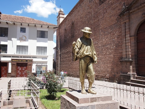 A bronze statue of a person wearing a traditional poncho and hat, standing on a stone pedestal. The statue is positioned in front of a historical stone building with arched doorways and adjacent to a modern office building with banners celebrating a milestone. The surrounding area includes small gardens and a wrought-iron fence.