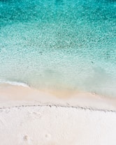 A stunning aerial view of a pristine beach with crystal-clear turquoise water gently lapping onto the soft, white sandy shore. The gradient of color from deep blue to light turquoise is visually striking, creating a serene and inviting atmosphere.