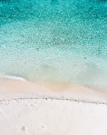 A stunning aerial view of a pristine beach with crystal-clear turquoise water gently lapping onto the soft, white sandy shore. The gradient of color from deep blue to light turquoise is visually striking, creating a serene and inviting atmosphere.