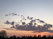 A scenic sunset with a silhouette of a couple walking hand-in-hand across a grassy field. The sky is filled with scattered clouds, with vibrant colors ranging from deep orange to pale blue.