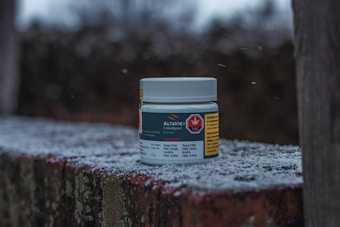 A white container with labels sits on a frost-covered, mossy brick surface. The background is blurred, with a muted color palette, suggesting a cold, outdoor environment.