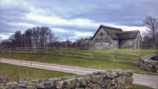 A rustic barn set in a countryside landscape, surrounded by a wooden fence and a neatly maintained grassy field. The sky is overcast, with clouds giving a moody atmosphere. A stone wall borders a gravel path leading towards the barn. Bare trees in the background suggest a late autumn or early winter setting.
