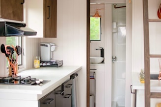 A compact and modern kitchen space is shown. There is a white countertop with a gas stove on the left, accompanied by a set of cooking utensils in copper containers. A coffee machine and a jar of spaghetti are also present. The cabinetry has a dark wood finish, and a small window provides natural light. In the background, a glimpse of a small, minimalistic bathroom with a sink and a shower can be seen.