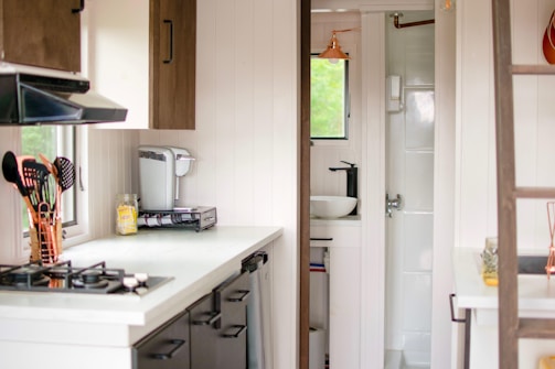 A compact and modern kitchen space is shown. There is a white countertop with a gas stove on the left, accompanied by a set of cooking utensils in copper containers. A coffee machine and a jar of spaghetti are also present. The cabinetry has a dark wood finish, and a small window provides natural light. In the background, a glimpse of a small, minimalistic bathroom with a sink and a shower can be seen.