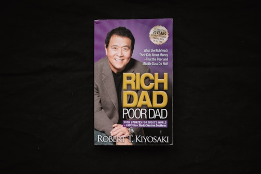A book cover is displayed against a dark background. The cover features a photograph of a smiling man in a suit alongside the title 'Rich Dad Poor Dad' in large yellow and white lettering. There is also a circular label indicating a 20th-anniversary edition.