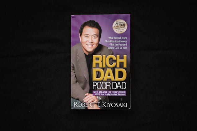 A book cover is displayed against a dark background. The cover features a photograph of a smiling man in a suit alongside the title 'Rich Dad Poor Dad' in large yellow and white lettering. There is also a circular label indicating a 20th-anniversary edition.