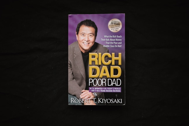 A book cover is displayed against a dark background. The cover features a photograph of a smiling man in a suit alongside the title 'Rich Dad Poor Dad' in large yellow and white lettering. There is also a circular label indicating a 20th-anniversary edition.