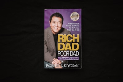 A book cover is displayed against a dark background. The cover features a photograph of a smiling man in a suit alongside the title 'Rich Dad Poor Dad' in large yellow and white lettering. There is also a circular label indicating a 20th-anniversary edition.