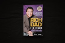 A book cover is displayed against a dark background. The cover features a photograph of a smiling man in a suit alongside the title 'Rich Dad Poor Dad' in large yellow and white lettering. There is also a circular label indicating a 20th-anniversary edition.