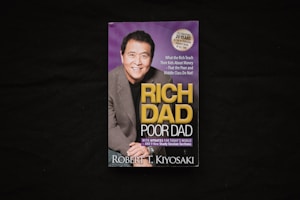 A book cover is displayed against a dark background. The cover features a photograph of a smiling man in a suit alongside the title 'Rich Dad Poor Dad' in large yellow and white lettering. There is also a circular label indicating a 20th-anniversary edition.