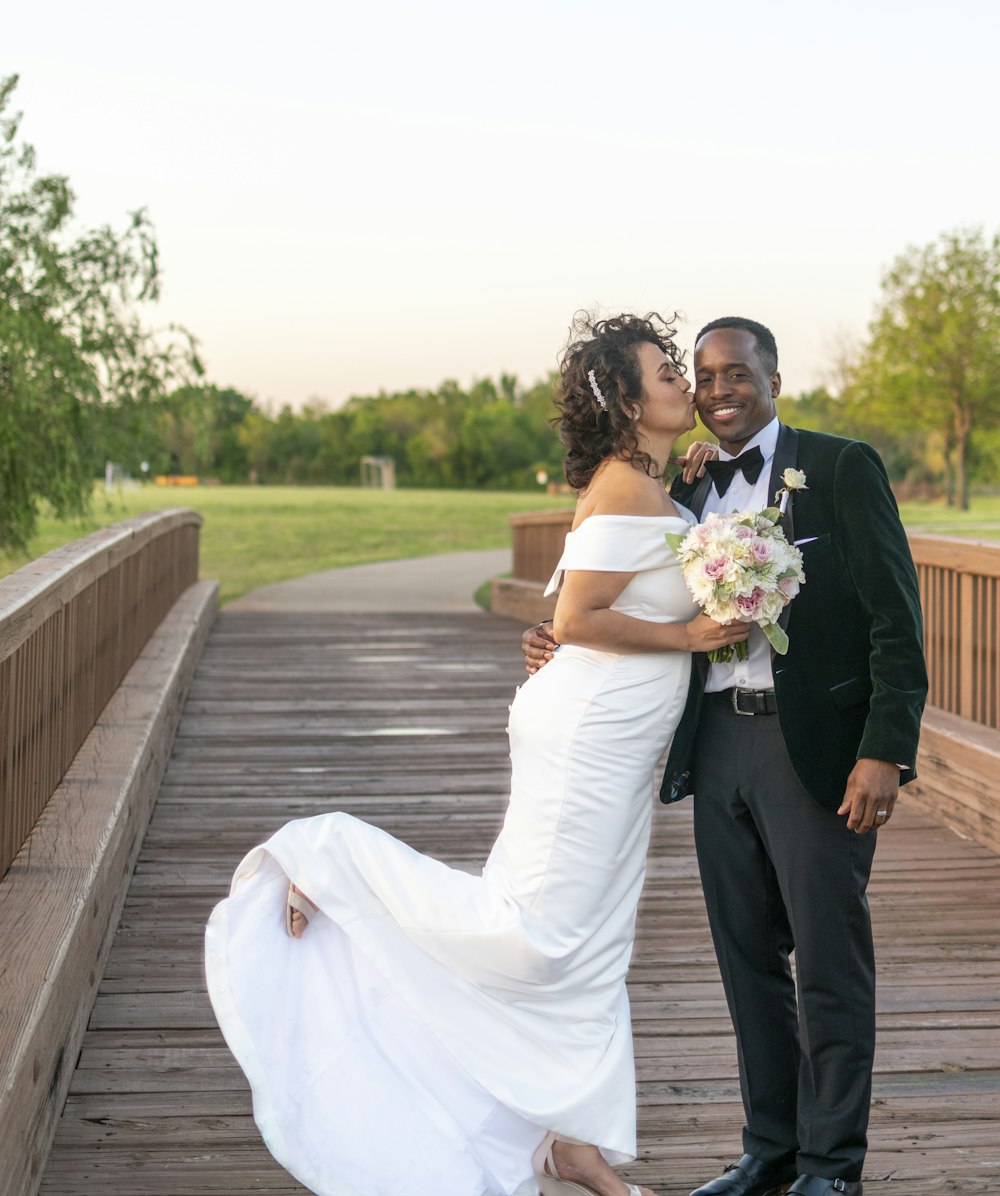 man and woman in wedding dress kissing on brown wooden bridge during daytime