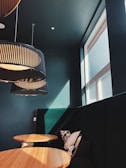 A modern interior with high ceilings features large, round wooden tables and dark colored upholstered seating. Large, black pendant lamps with ribbed designs hang from the ceiling, casting soft shadows. Sunlight streams through a high window, creating a warm and inviting atmosphere. A few decorative pillows with minimalist patterns are placed on the bench seats.