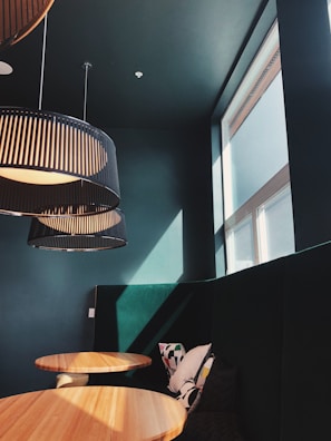 A modern interior with high ceilings features large, round wooden tables and dark colored upholstered seating. Large, black pendant lamps with ribbed designs hang from the ceiling, casting soft shadows. Sunlight streams through a high window, creating a warm and inviting atmosphere. A few decorative pillows with minimalist patterns are placed on the bench seats.