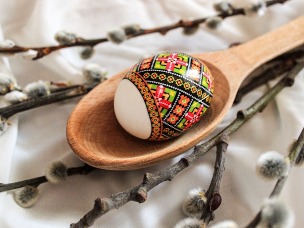 A wooden spoon holds a beautifully decorated Easter egg with intricate, colorful patterns featuring red, green, and black designs. The spoon and egg are placed on a soft white fabric surrounded by pussy willow branches, which have fuzzy, grey catkins.
