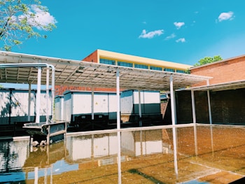 A partially covered industrial area features reflective water on the ground, with a backdrop of buildings including a structure with yellow and red accents. There are several utility cabins or containers and a large tub-like structure. Clear blue sky with some scattered clouds and a tree branch are visible.
